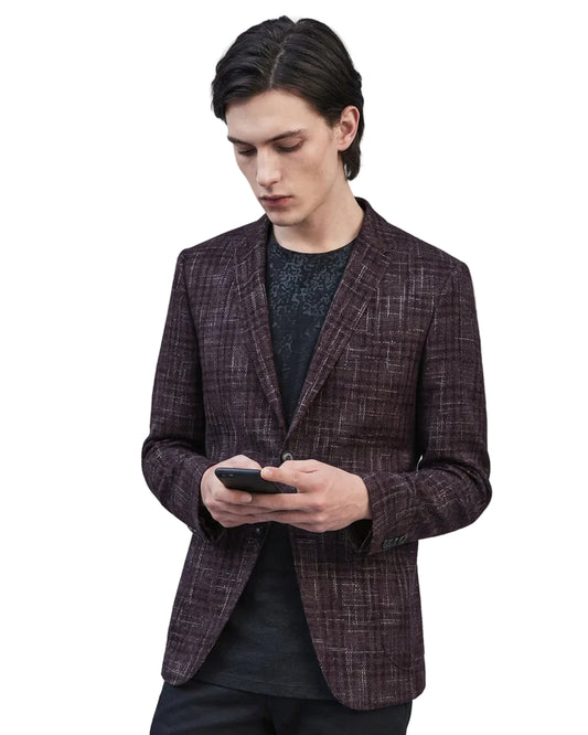 A man wearing a dark brown and black plaid blazer over a black textured shirt, holding a black smartphone against a dark gray background.